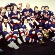 Skupna fotografija hokejaške reprezentacije SAD-a iz 1980. godine, poznate kao "Miracle on Ice" generacija. Igrači u bijelim dresovima s natpisom "USA" i plavim kacigama stoje i sjede u nekoliko redova na ledu dvorane u Lake Placidu. U sredini se ističu trener Herb Brooks i igrači poput Mikea Eruzionea i Marka Pavelicha. Fotografija je u retro stilu, zabilježena nakon osvajanja zlatne olimpijske medalje.