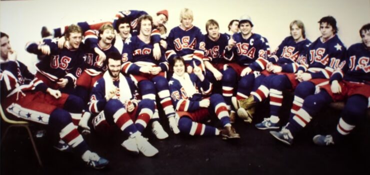 Skupna fotografija hokejaške reprezentacije SAD-a iz 1980. godine, poznate kao "Miracle on Ice" generacija. Igrači u bijelim dresovima s natpisom "USA" i plavim kacigama stoje i sjede u nekoliko redova na ledu dvorane u Lake Placidu. U sredini se ističu trener Herb Brooks i igrači poput Mikea Eruzionea i Marka Pavelicha. Fotografija je u retro stilu, zabilježena nakon osvajanja zlatne olimpijske medalje.