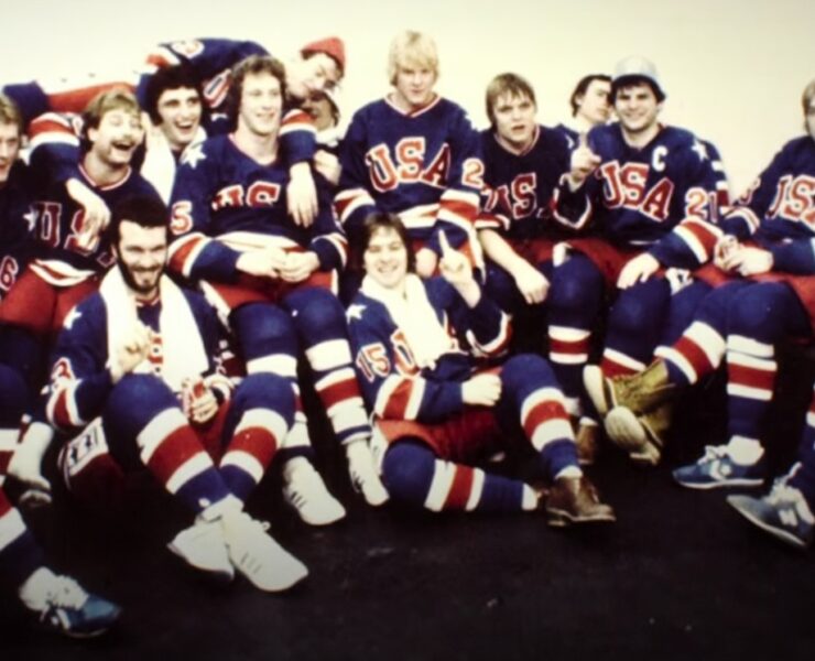 Skupna fotografija hokejaške reprezentacije SAD-a iz 1980. godine, poznate kao "Miracle on Ice" generacija. Igrači u bijelim dresovima s natpisom "USA" i plavim kacigama stoje i sjede u nekoliko redova na ledu dvorane u Lake Placidu. U sredini se ističu trener Herb Brooks i igrači poput Mikea Eruzionea i Marka Pavelicha. Fotografija je u retro stilu, zabilježena nakon osvajanja zlatne olimpijske medalje.