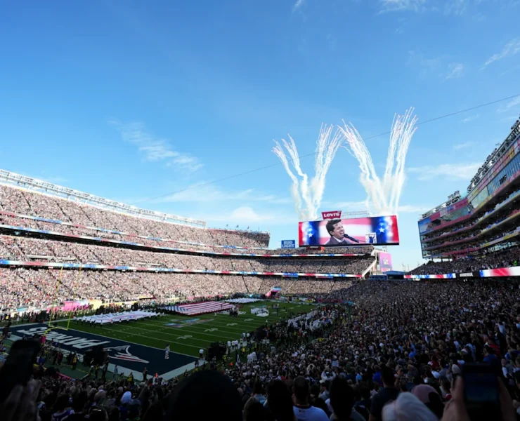 Široki kadar prepunog Levi's stadiona tijekom ceremonije prije početka Super Bowla. Na velikom ekranu prikazan je pjevač, dok se iza ekrana u nebo diže bijeli vatromet. Na terenu su postavljene velike američke zastave, a tribine su ispunjene tisućama gledatelja pod vedrim plavim nebom.